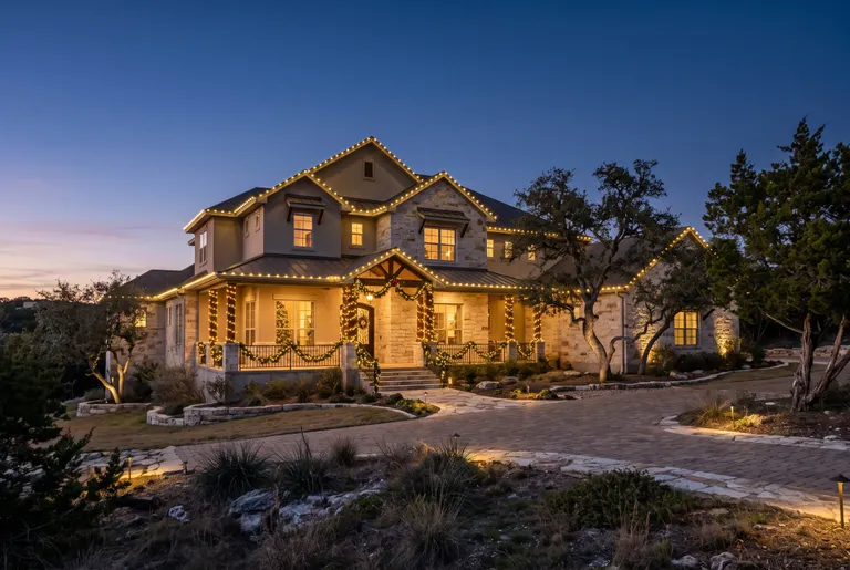 Two-story stone and stucco home with a layered lit roofline, wrapped porch columns and lit garland at the entry