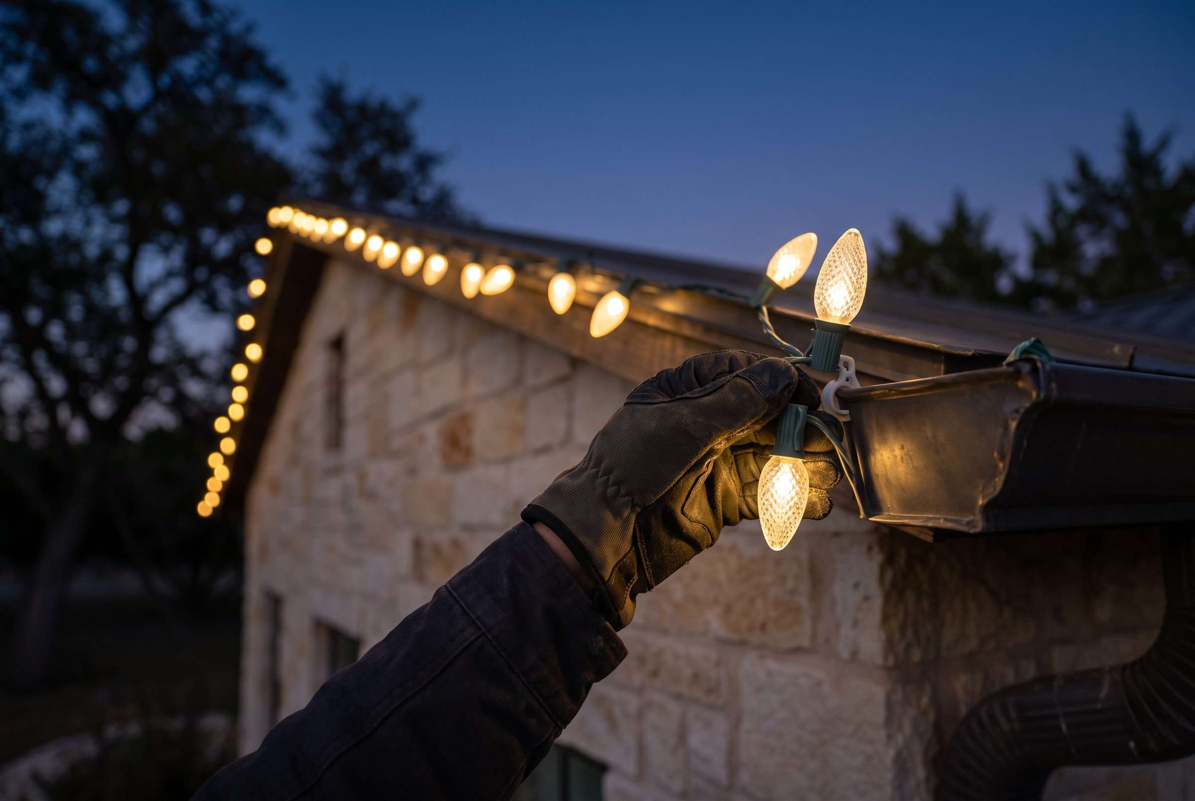 Gloved hand clipping a warm white C9 light strand to a gutter at dusk