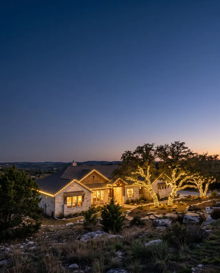 Single-story limestone ranch home on a Hill Country hillside at blue hour with warm white C9 bulbs on the roofline and two live oaks wrapped in lights