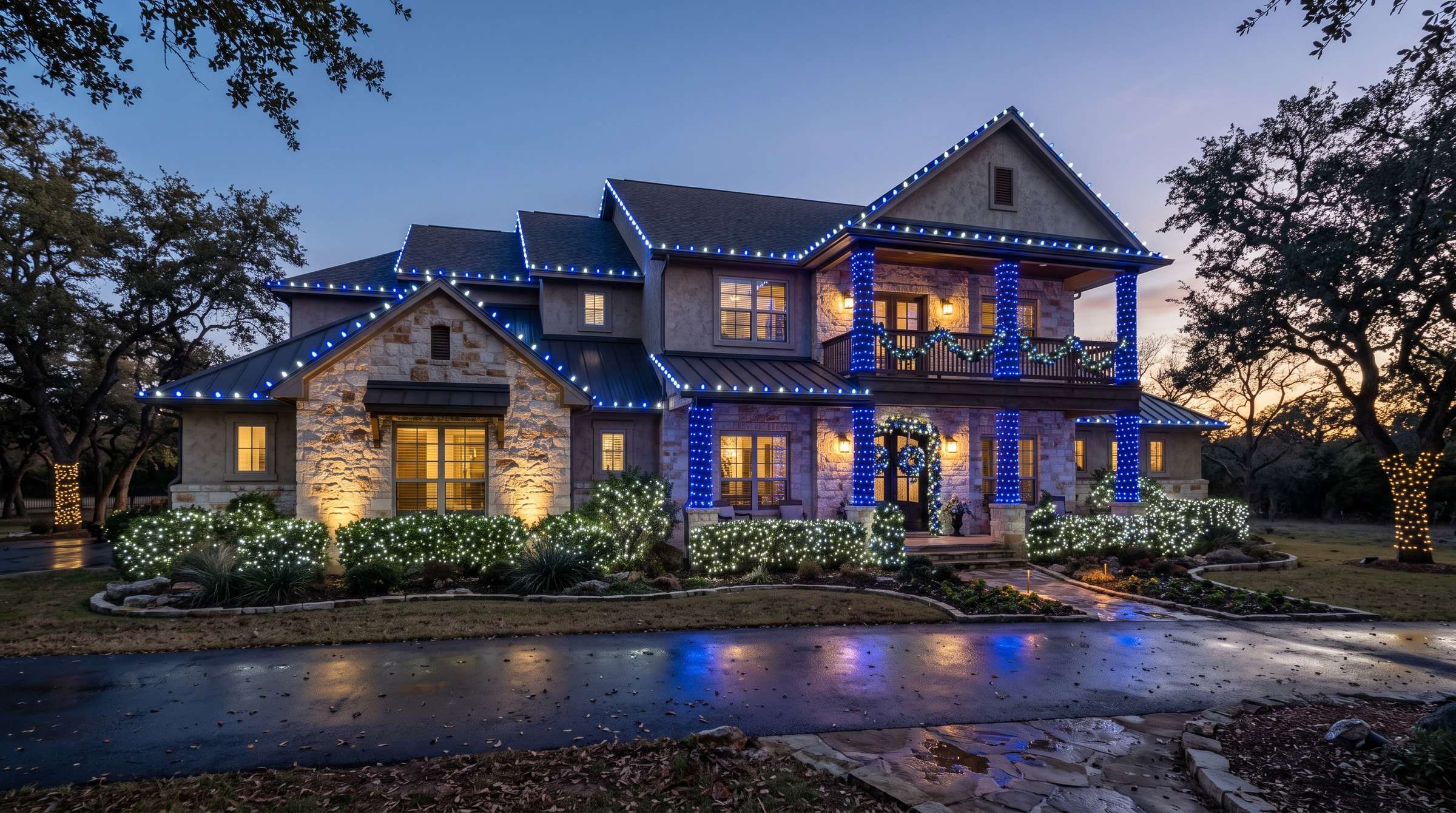 Two-story stone home with a lit roofline, wrapped shrubs and trees, and a lit wreath and garland at the door